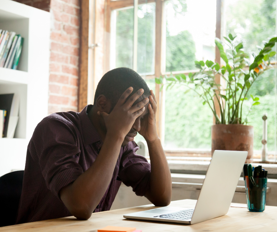 Man of color sitting at a desk with both hands on his head looking at a laptop.